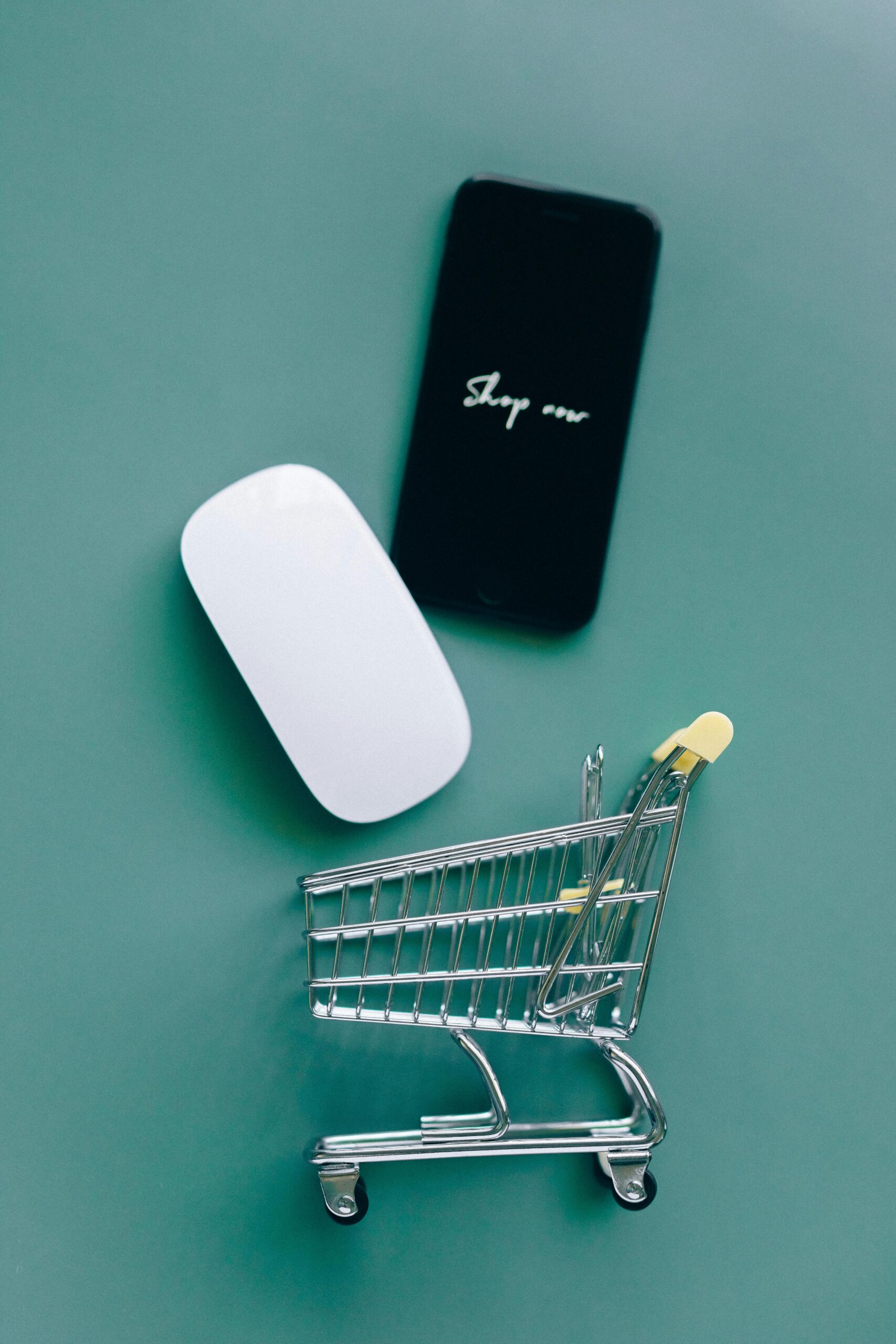 Flat lay of a miniature shopping cart, smartphone, and computer mouse on teal background.